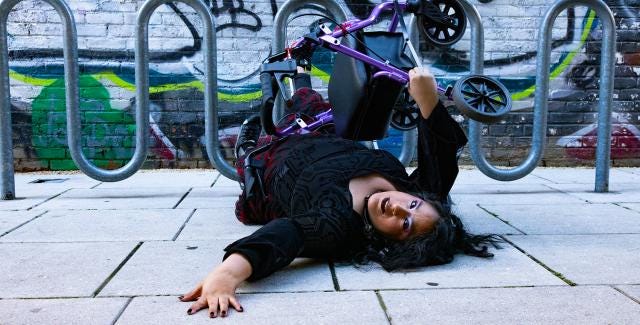 A photo of Vanessa, who is a Chicana woman with caramel skin is lying down on the concrete floor with her arm reaching out as she is looking back towards the camera. Her walker Pluto is balancing on top of her. She is wearing a gothic long sleeve blouse with a flowered black and red skirt. In the background is a looping metal bicycle stand with a graffiti filled wall.