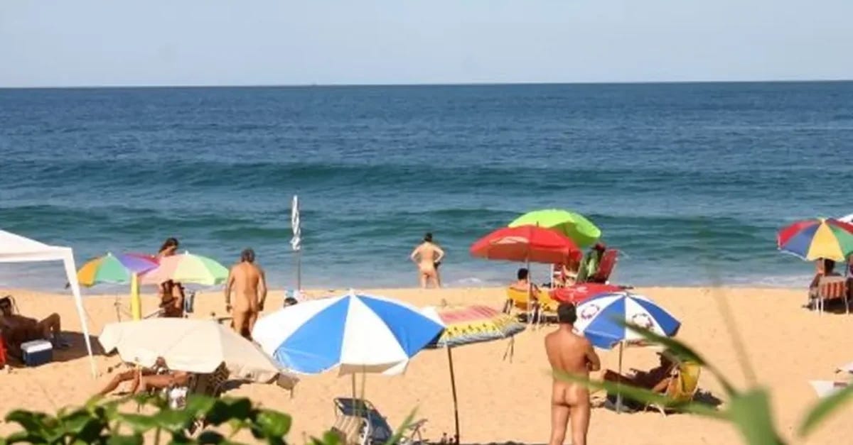 Wide view of Praia do Pinho with beachgoers spread across the sand, some nude and others seated under colorful umbrellas, facing the ocean on a clear day in Santa Catarina, Brazil.