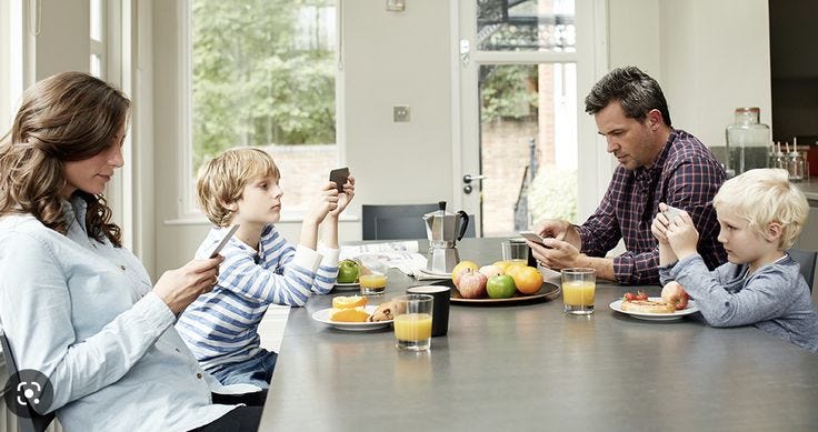 This may contain: a family sitting around a kitchen table with food on the plates and looking at their phones This may contain: a family sitting around a kitchen table with food on the plates and looking at their phones