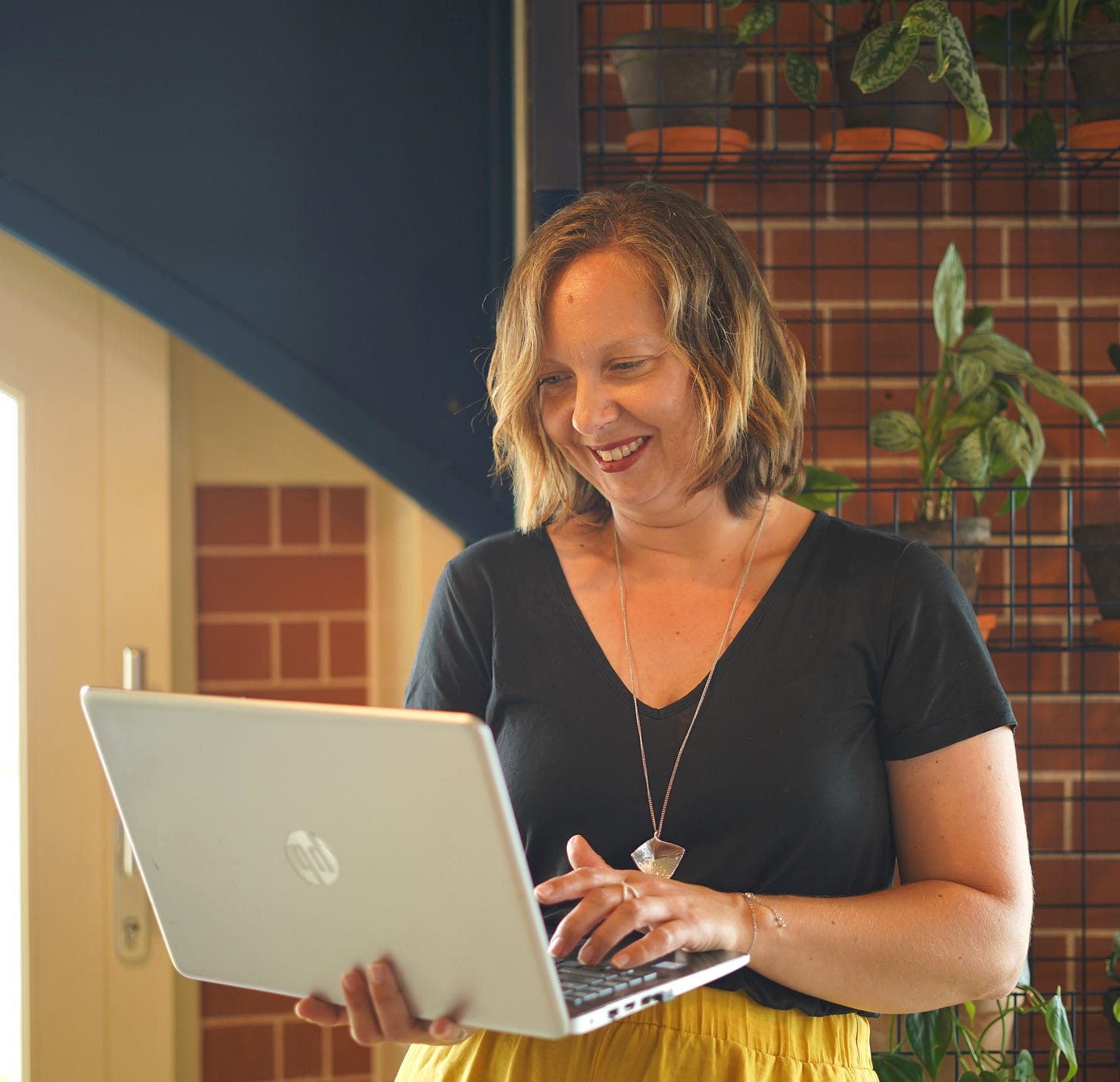 Allegra Chapman smiling while working on a laptop indoors, standing in front of shelves filled with houseplants and warm-toned brick walls. She wears a black top, yellow skirt, and silver pendant necklace in a relaxed creative workspace