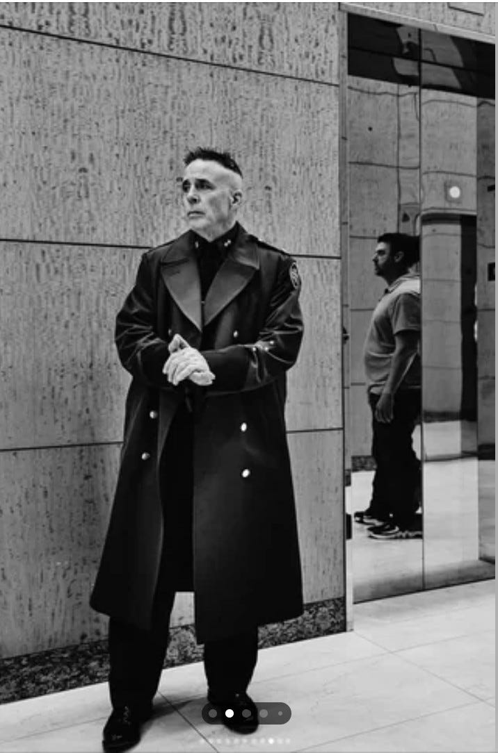 Man standing in front of building, wearting long black leather coat and haircut reminiscent of the Nazi SS fashion