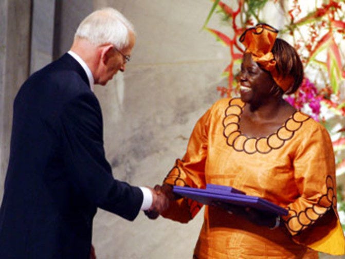 Wangari Maathai, dressed in a vibrant orange traditional African outfit with black detailing and a matching headwrap, smiles as she receives the Nobel Peace Prize. She is shaking hands with a white-haired man in a dark suit, while holding a purple award folder. Behind them, a floral arrangement adds color to the setting. The moment captures Maathai’s historic achievement as the first African woman to receive the Nobel Peace Prize. Wangari Maathai, dressed in a vibrant orange traditional African outfit with black detailing and a matching headwrap, smiles as she receives the Nobel Peace Prize. She is shaking hands with a white-haired man in a dark suit, while holding a purple award folder. Behind them, a floral arrangement adds color to the setting. The moment captures Maathai’s historic achievement as the first African woman to receive the Nobel Peace Prize.