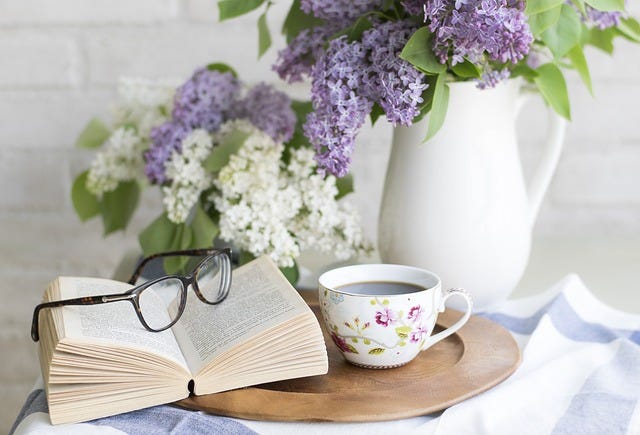 Photo of open book with glasses on top. Behind are flowers in a vase and to the side a teacup with tea. Photo of open book with glasses on top. Behind are flowers in a vase and to the side a teacup with tea.