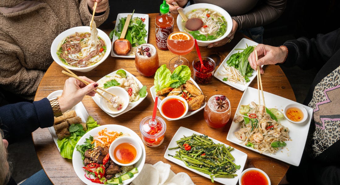 A table filled with pho, vegetables, and cocktails, with people sat around it