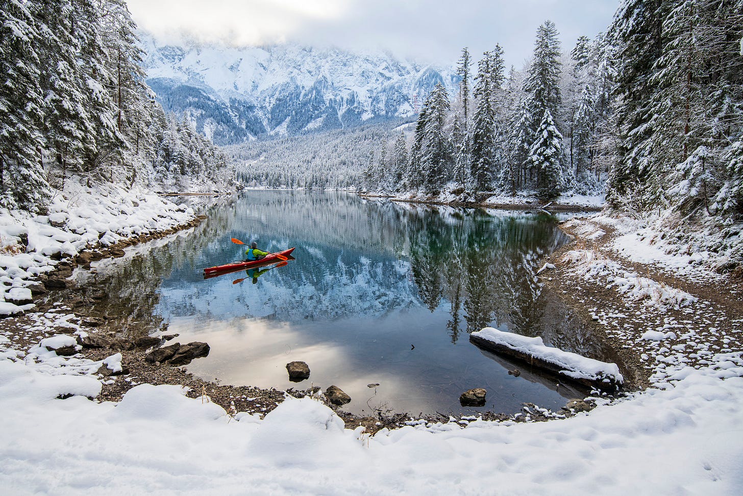 The Eibsee in winter surrounded by snow. There is a canoeist on the lake and behind are the snow-covered Wetterstein mountains