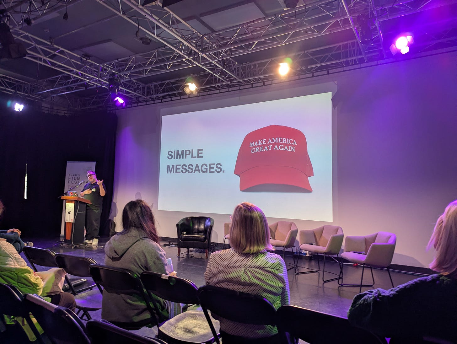 Man standing to left of a screen with the words Simle Messages and a red cap with Make America Great Again written on it. Man standing to left of a screen with the words Simle Messages and a red cap with Make America Great Again written on it.