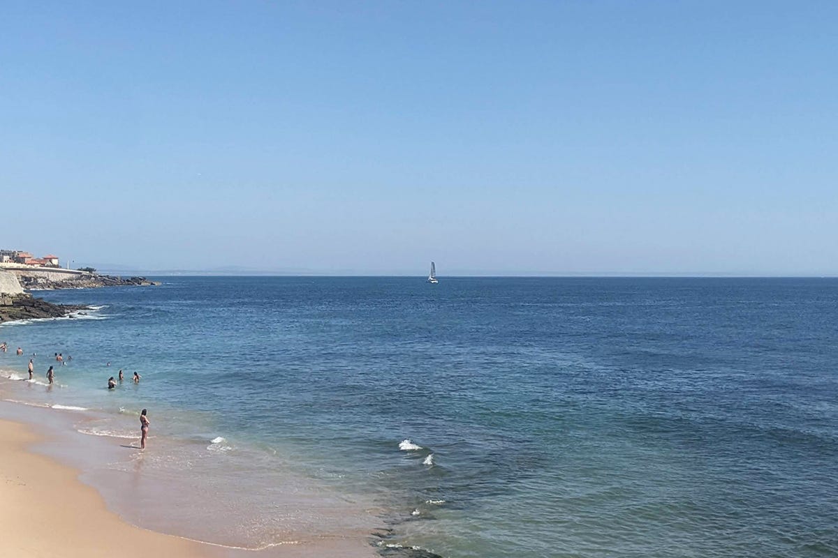 photo of a sunny beachscape in portugal. there are people standing and wading in the shallow parts of the water and a sailboat in the distance. photo of a sunny beachscape in portugal. there are people standing and wading in the shallow parts of the water and a sailboat in the distance.