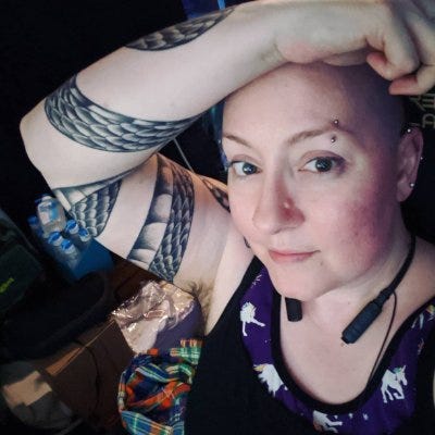 a person with pale skin and an arm tattoo, smiling at the camera with their right arm resting above their head