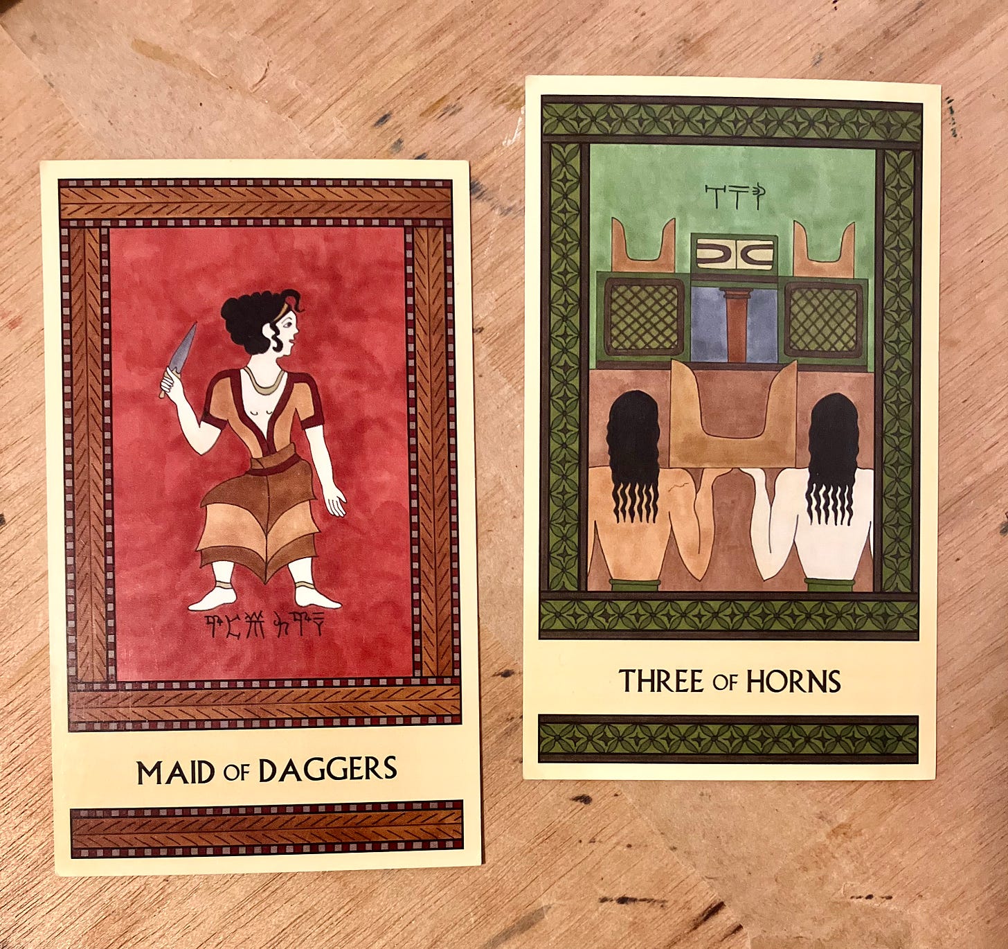 Two Minoan Tarot cards side by side on a paint-spattered wooden surface (my art desk).  The Maid of Daggers is painted in shades of deep red and brown. It shows a Minoan girl standing, her head turned to the right, holding up a dagger in the opposite hand. The Three of Horns is painted in shades of deep green and brown. It shows two people from the back, from the waist up, carrying a large pair of sacred horns between them. They are facing a shrine that already contains two pairs of sacred horns. The implication is that they're working together to put the third pair of horns on the altar. 