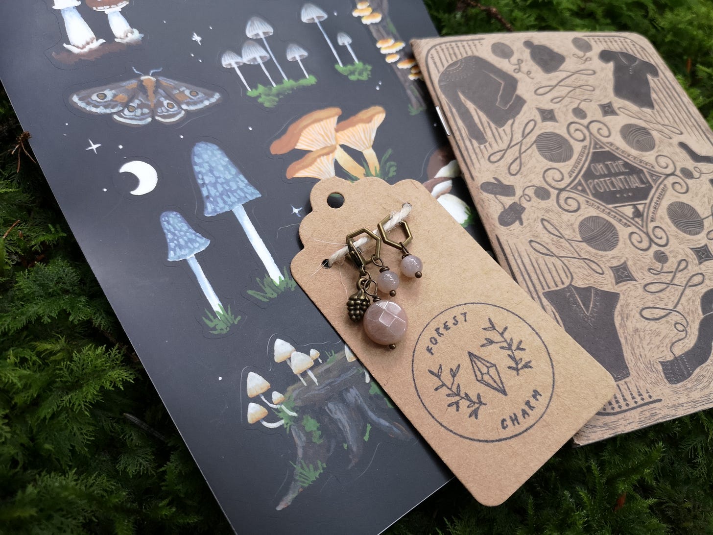 Image description: A close up photo of a gift bundle sitting on top of some squishy moss. On top is a beautiful card of handmade stitch markers made with moonstone beads and metal charms. Beneath these are some mushroom stickers and a knitting notebook.