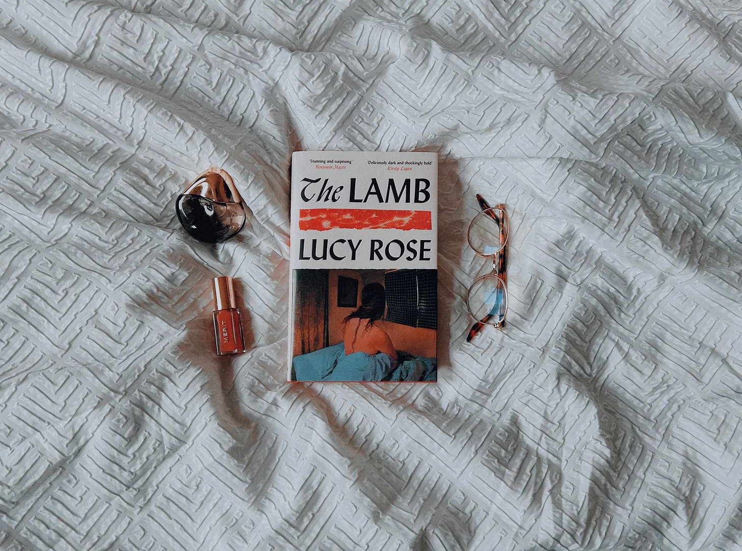the lamb (novel) laid out on a clean but crumpled blankets. perfume, lipstick and a pair of glasses neatly strewn close by. 