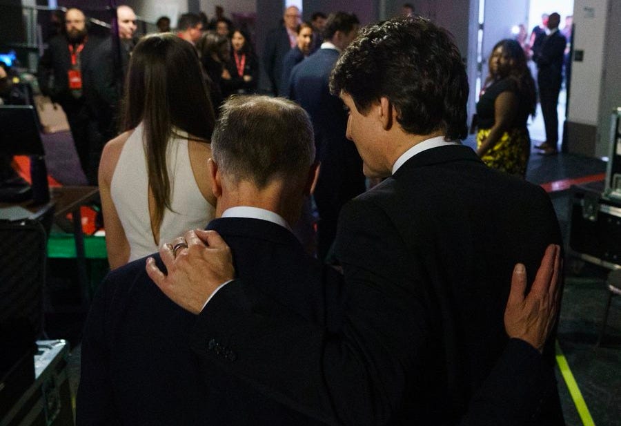 The image captures a moment of camaraderie between two individuals, one of whom is likely Prime Minister Justin Trudeau, as suggested by the post text from Mark Carney thanking him for his leadership and service to Canada. The setting appears to be a formal event or gathering, with several people in the background dressed in formal attire, indicating a significant occasion. The two men are seen from behind, with one placing his hand on the other's shoulder, symbolizing support and friendship. The composition focuses on this gesture, emphasizing the personal connection and mutual respect between them. The lighting and clarity of the image suggest it was taken in a well-lit indoor environment, possibly a conference or a political event.