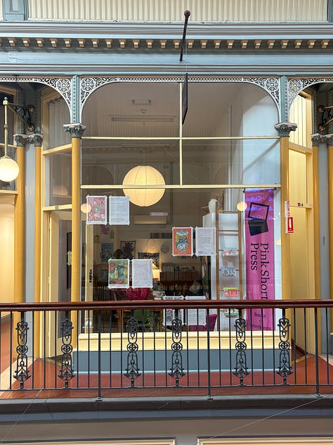A stack of wrapped books, a promotion for the 'At Our Mothers' Table' reading event on 2 August, and the front of the Pink Shorts Press shop in Adelaide Arcade.