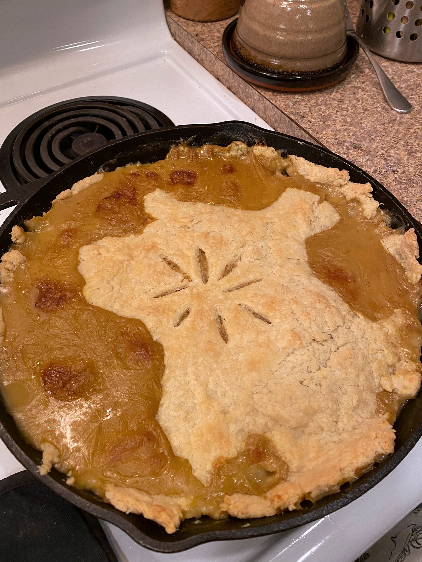 A cast iron pan with a pot pie, an asterisk shape scored in the centre. A brownish curried gravy has spilled overtop of the crust in several places. A cast iron pan with a pot pie, an asterisk shape scored in the centre. A brownish curried gravy has spilled overtop of the crust in several places.