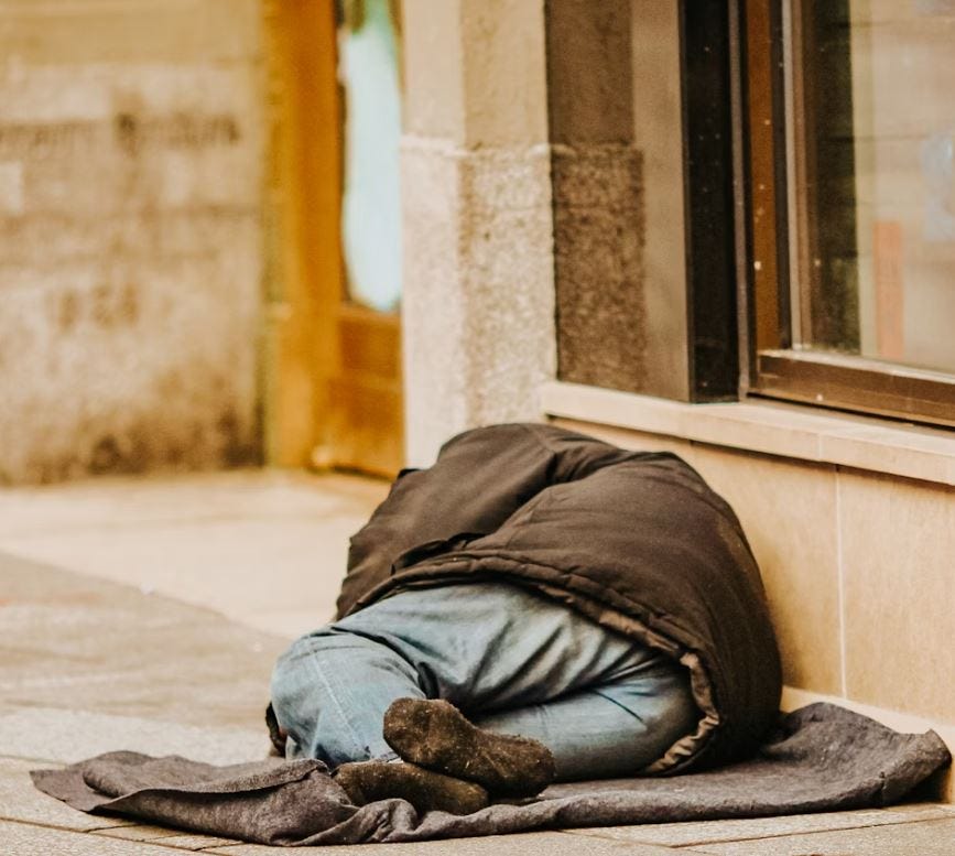 A person experiencing homelessness sleeps on the sidewalk, wrapped in a thick jacket, illustrating the harsh realities of co-occurring substance abuse and mental illness.