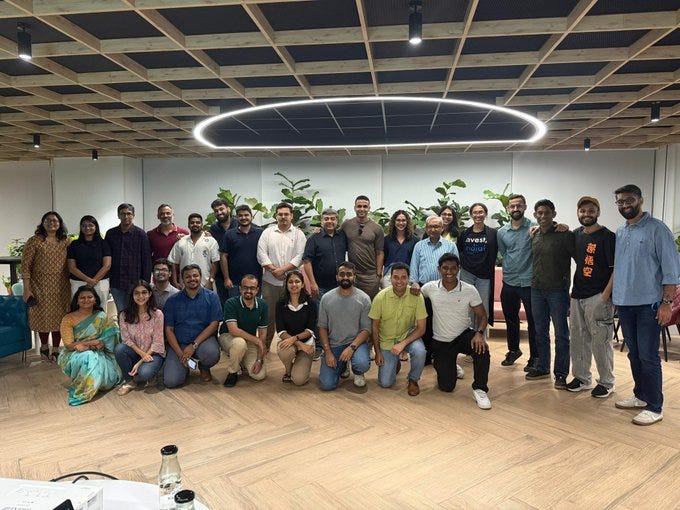 A group photo of Nithin Kamath, Manoj Malgharia, and others from Rainmatter and SocialAlpha. They are standing and kneeling in a modern office with wooden flooring, a circular ceiling light, and green plants. Some individuals wear casual attire like jeans and t-shirts, while others wear traditional clothing like sarees.