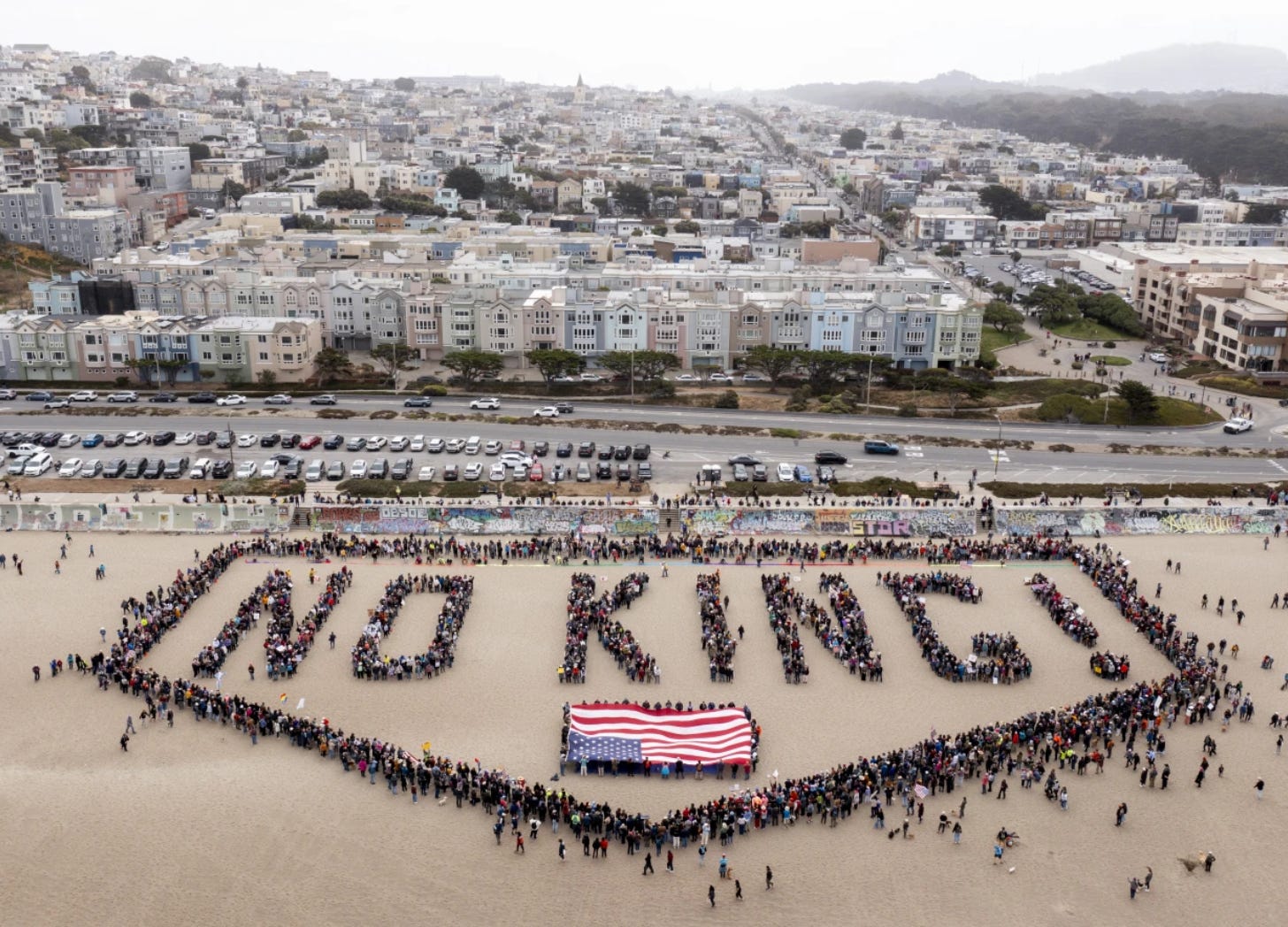 Aerial photo of a large group of people gathered on Ocean Beach in San Francisco, forming the giant phrase “NO KING!” with their bodies, surrounded by a border of additional people. Below the words, a large, inverted American flag is laid out on the sand. In the background are pastel-colored row houses, cars, and a foggy urban skyline. Aerial photo of a large group of people gathered on Ocean Beach in San Francisco, forming the giant phrase “NO KING!” with their bodies, surrounded by a border of additional people. Below the words, a large, inverted American flag is laid out on the sand. In the background are pastel-colored row houses, cars, and a foggy urban skyline.