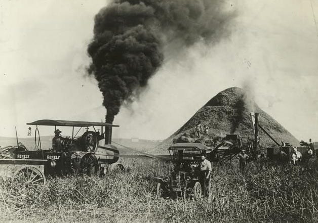 A thresher in a field of grass with a small vehicle next to it, in black and white, some time in the 1800s