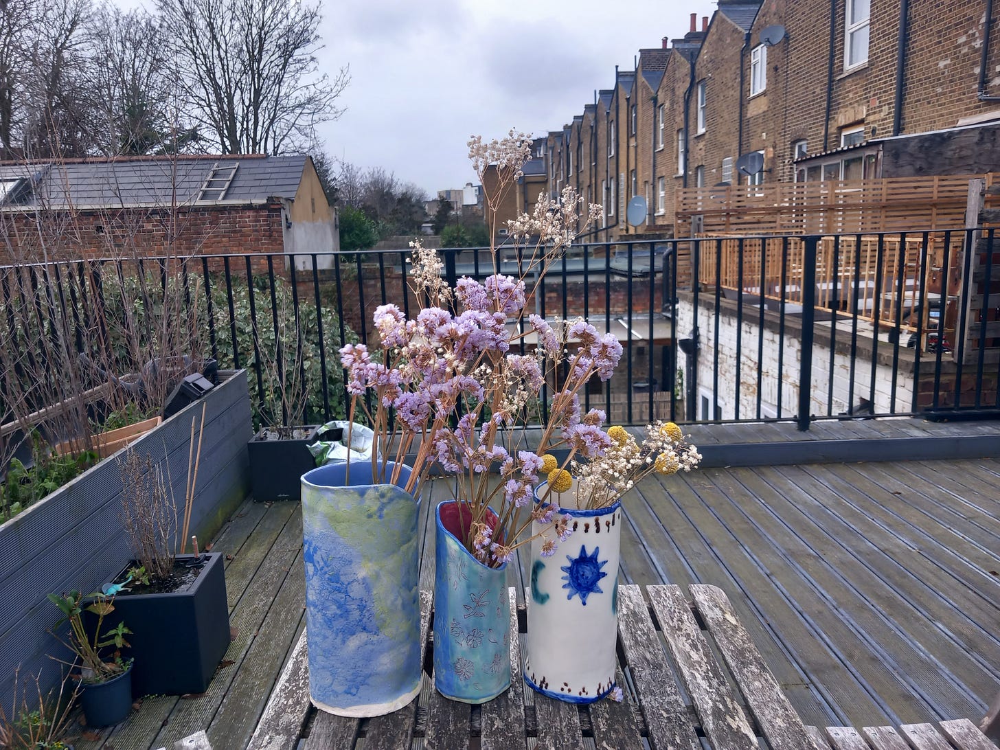 A blue and yellow vase, a green and blue vase with pink illustrations of shells and a white vase with a green moon and blue star on a rooftop patio