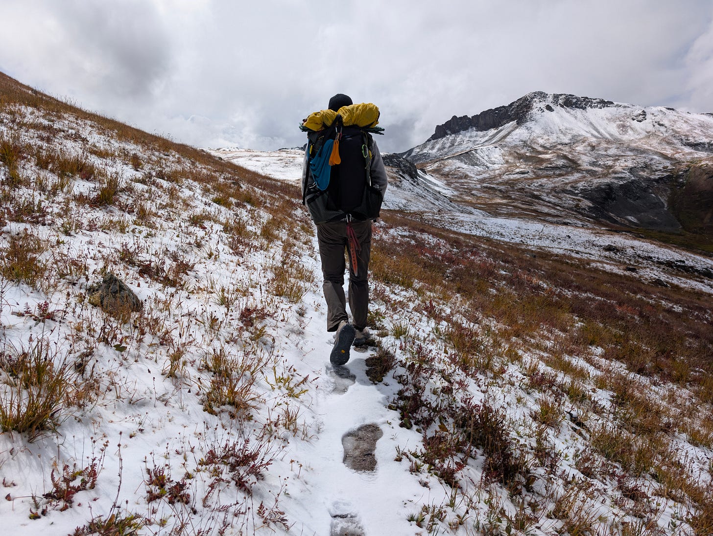 Backpacker walking uphill through early snowfall in alpine terrain, carrying a full pack across high-elevation tundra.