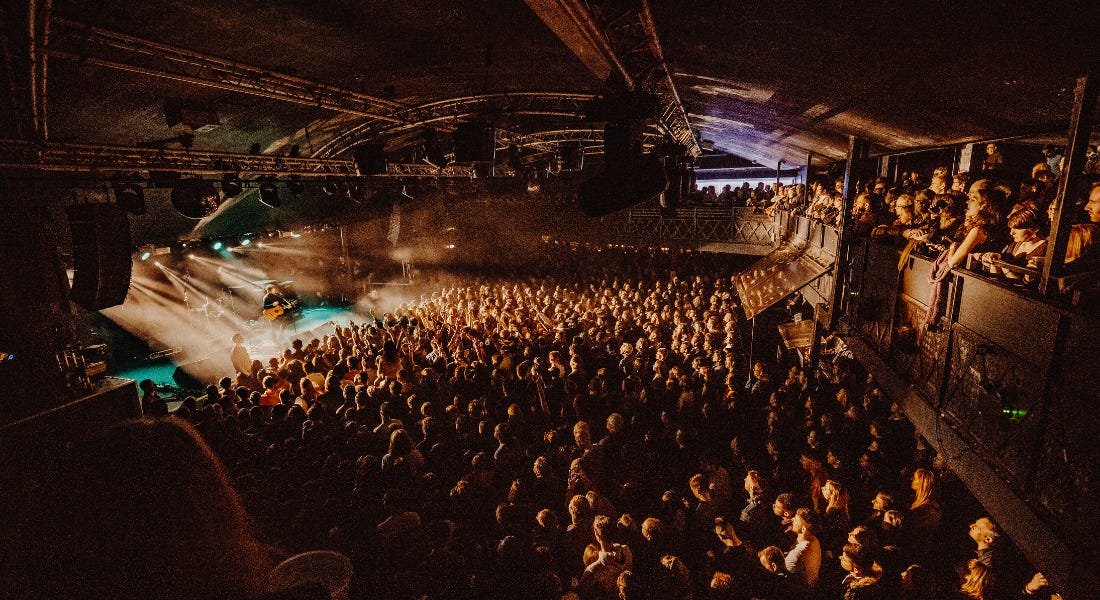 A dark concert venue with a large crowd at the bottom and on the balcony, and a blue stage to the right