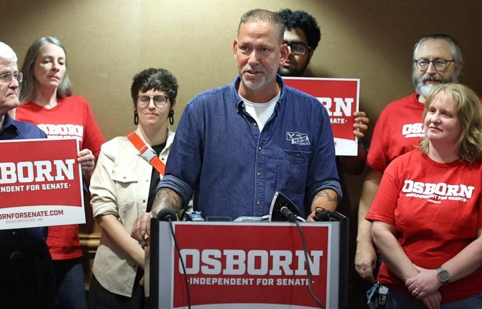 Dan Osborn, wearing a blue work shirt, speaks at a lectern as supporters gather around him