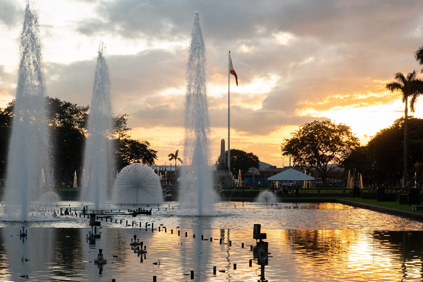 Reflecting pool and water fountains at Rizal Park in Manila in the evening before sunset. The sun is painting the clouds golden and yellow, the colors reflecting in the water.