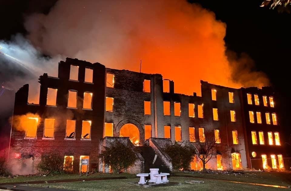 Color photo of a mostly-gutted 4-story brick building still burning at night, the sky above it glowing read from the flames Color photo of a mostly-gutted 4-story brick building still burning at night, the sky above it glowing read from the flames