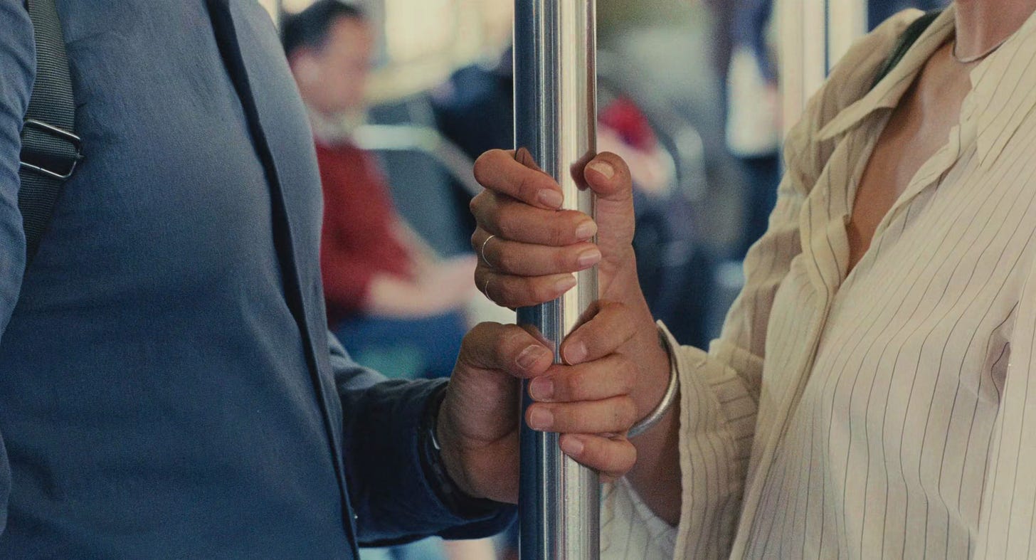 Close nas mãos de Nora e Hae Sung próximas em uma barra de apoio do metrô, quase se tocando mas sem estabelecer contato, simbolizando a proximidade e a distância entre eles.