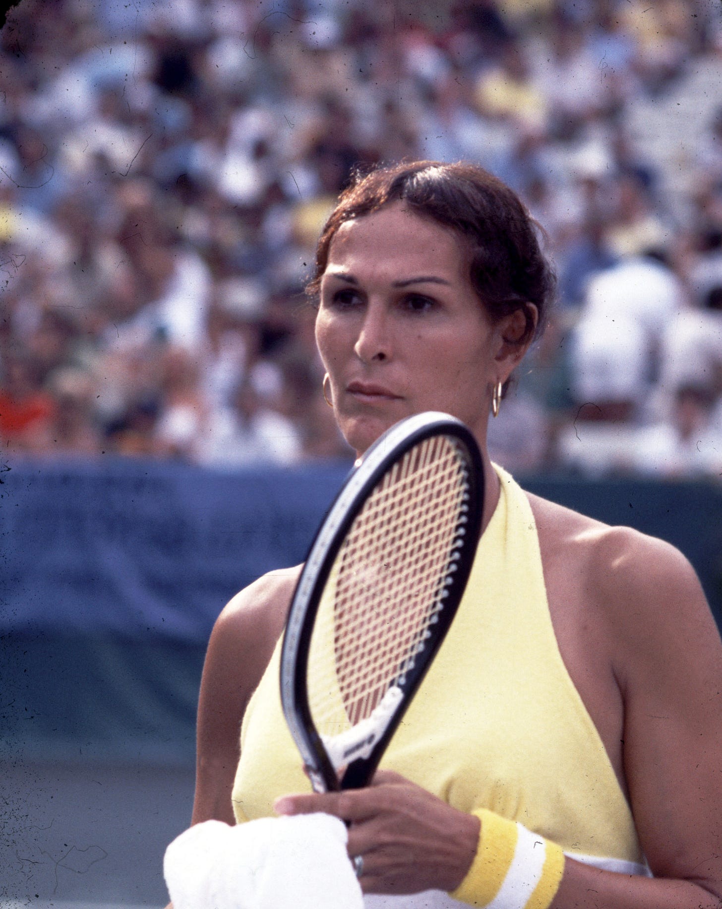 Rene Richards, on a tennis court, holding her racket with fans blurred in the background