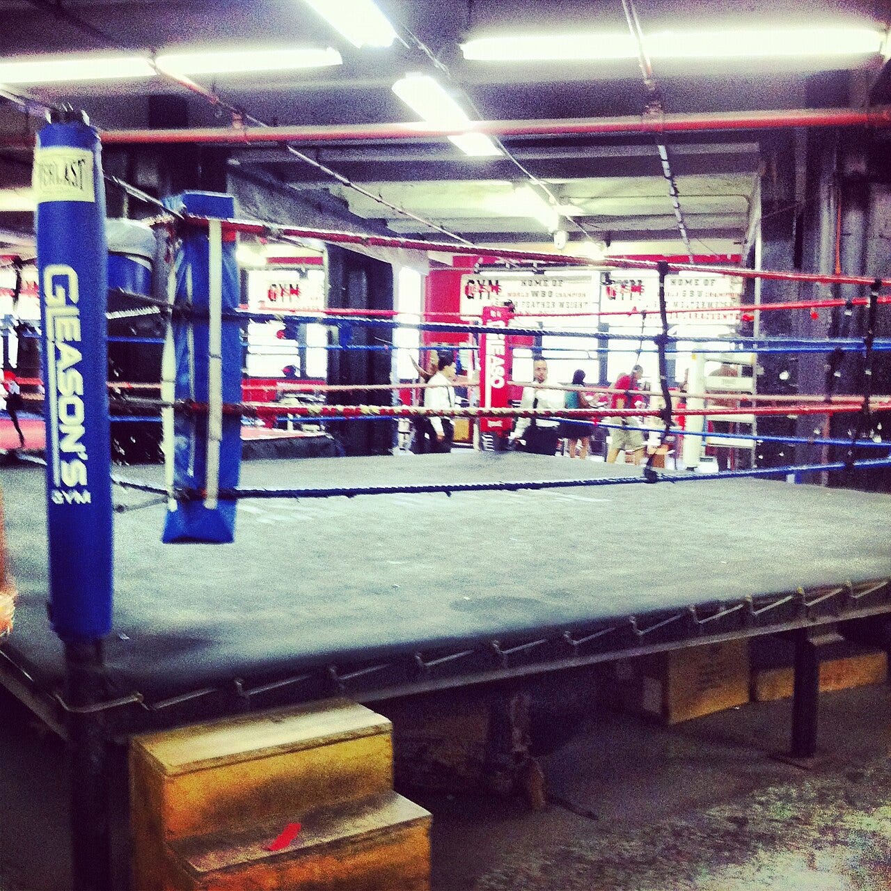 A boxing ring with a blue Gleason's Gym corner pad in the foreground and red and blue ropes, inside a brightly lit gym with banners on the far wall