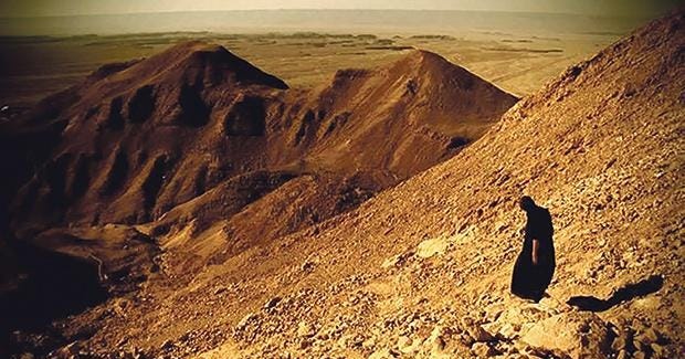 A lone figure in black ascends a steep, rocky desert slope under harsh sunlight—small against the vast, unforgiving terrain. The scene evokes ascetic pilgrimage, penance, and the silent weight of vocation.
