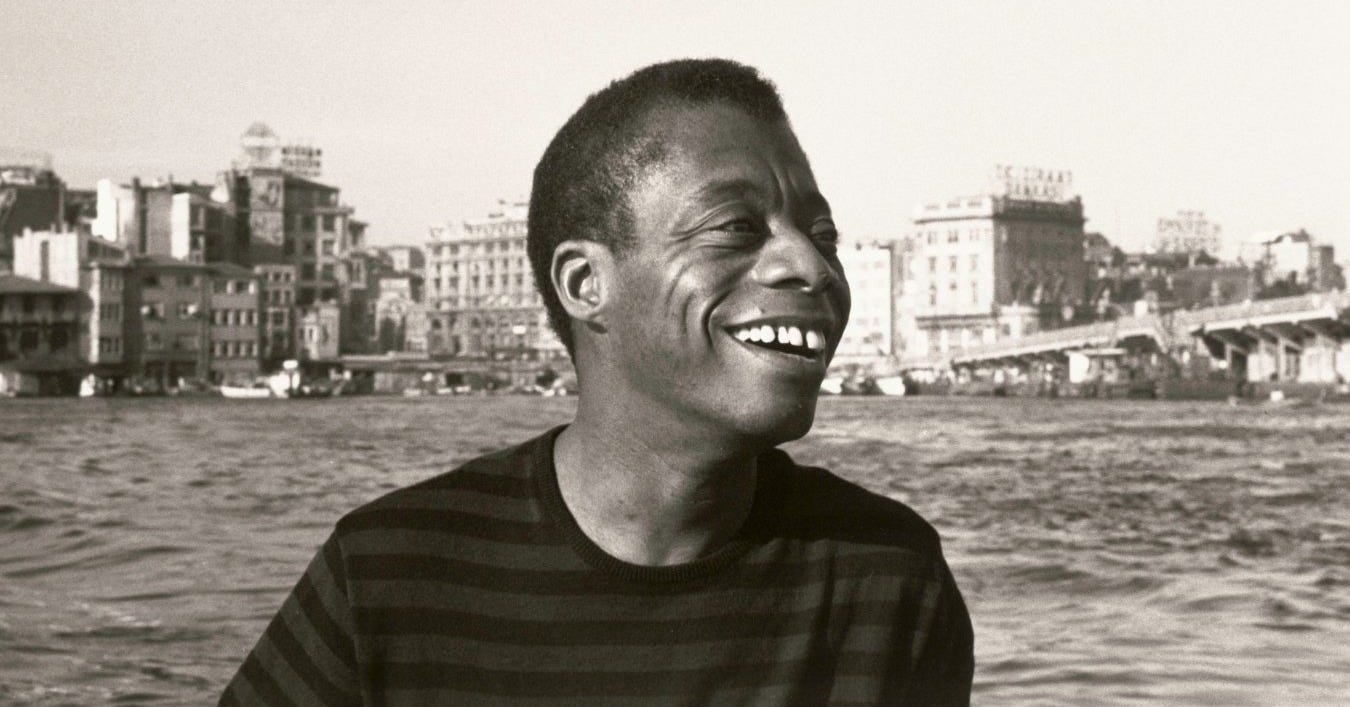 A black-and-white photograph of James Baldwin in a dark shirt. He is on the water of the Golden Horn.