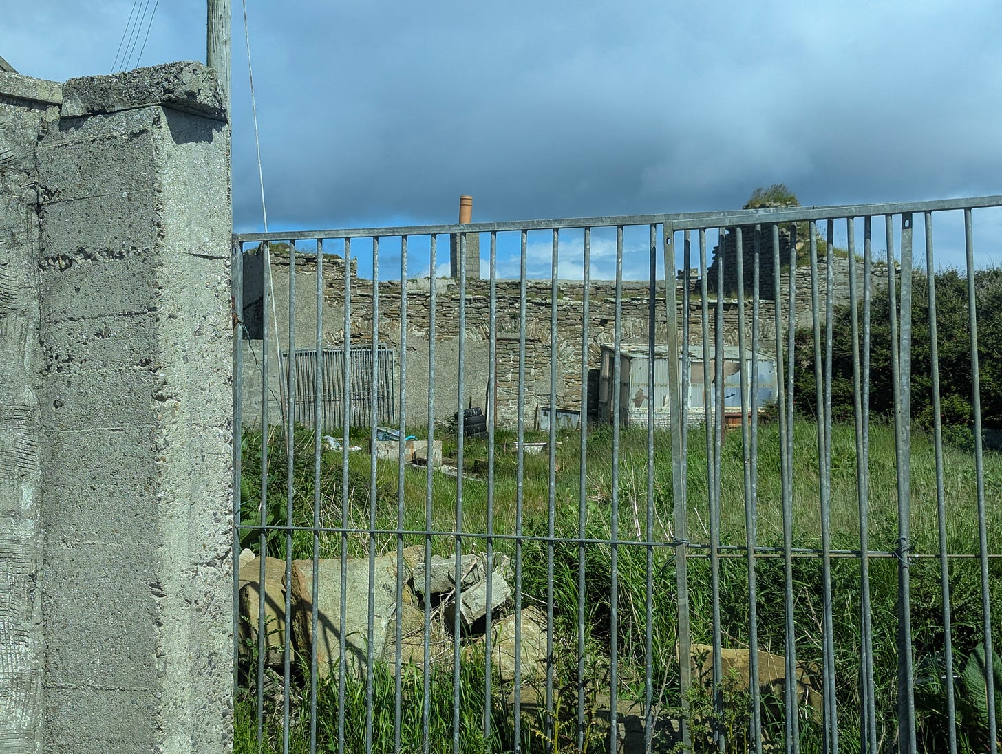 through a metal fence, we see a disused yard where a ruin on the opposite side has bricked-up archways in the stone wall.