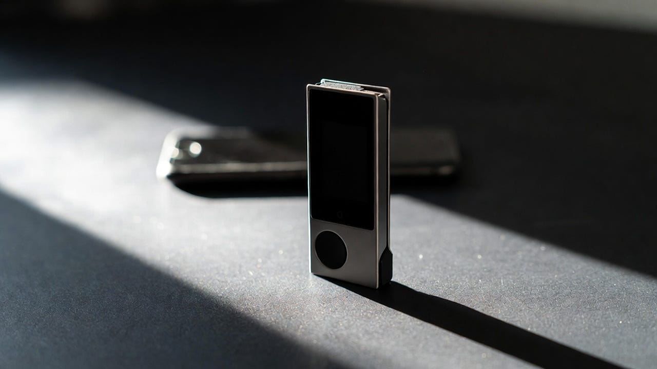 A Ledger hardware wallet stands upright on a dark surface under dramatic directional light, with a smartphone lying flat and powerless in the background, representing the recommendation to move cryptocurrency off vulnerable mobile devices onto offline hardware storage
