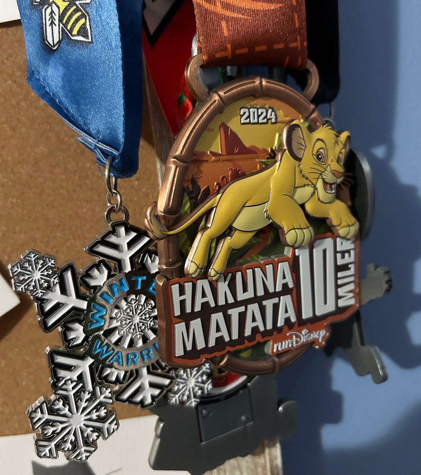 Photo of race medals hanging on a cork board. Most prominent are a blue and white snowflake "Winter Warriors" medal and an orange and gold Disney-themed "Hakuna Matata" medal.