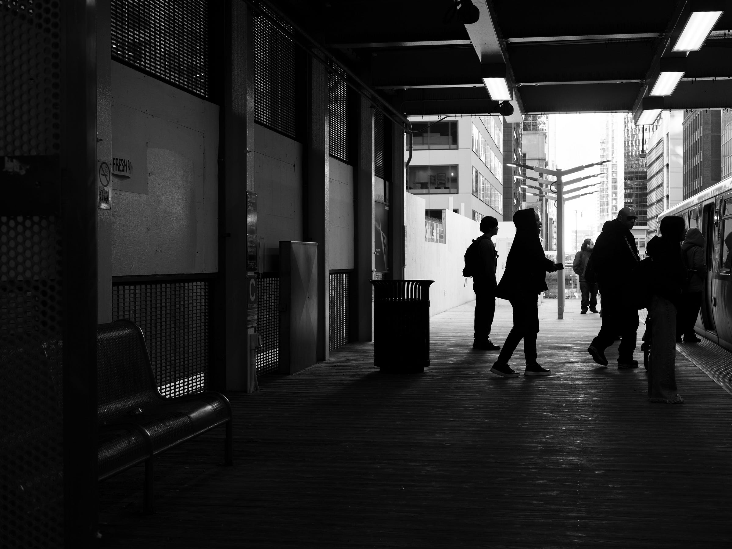 Black and white photograph of a Chicago train platform, where silhouetted commuters board a train. The overhead lights cast a glow on the wooden platform, contrasting with the bright city skyline in the background. A lone bench sits empty to the side, adding to the stillness of the moment. Black and white photograph of a Chicago train platform, where silhouetted commuters board a train. The overhead lights cast a glow on the wooden platform, contrasting with the bright city skyline in the background. A lone bench sits empty to the side, adding to the stillness of the moment.