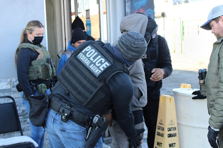 Person with vest labeled 'Police Federal Agent' shown patting down person in jeans and hoodie as other people stand and watch Person with vest labeled 'Police Federal Agent' shown patting down person in jeans and hoodie as other people stand and watch
