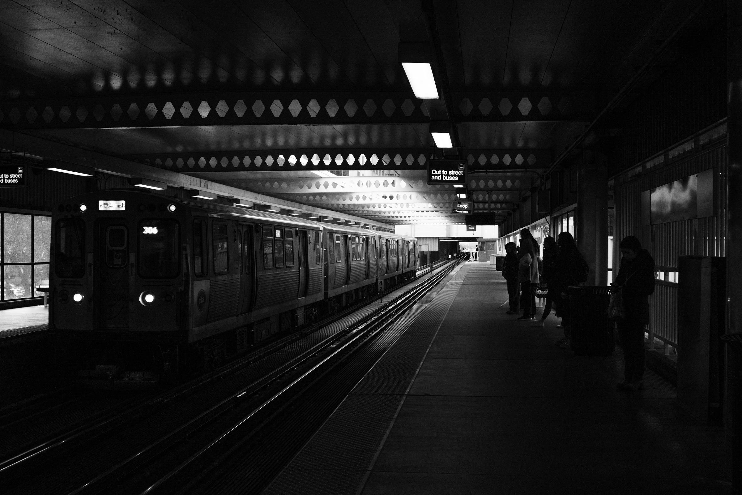 Black and white photo of a train arriving at an elevated station, with passengers waiting on the platform under dim overhead lights. Black and white photo of a train arriving at an elevated station, with passengers waiting on the platform under dim overhead lights.