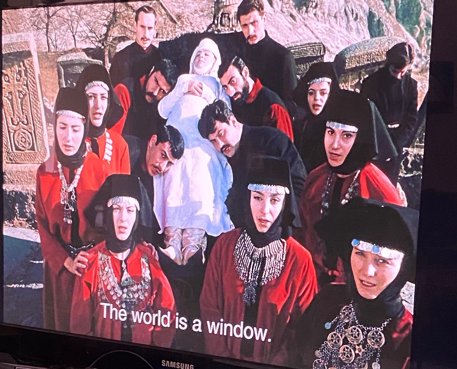 A group of women dressed in red and a group of men dressed in black carry a woman in a white dress. The caption reads: The world is a window."
