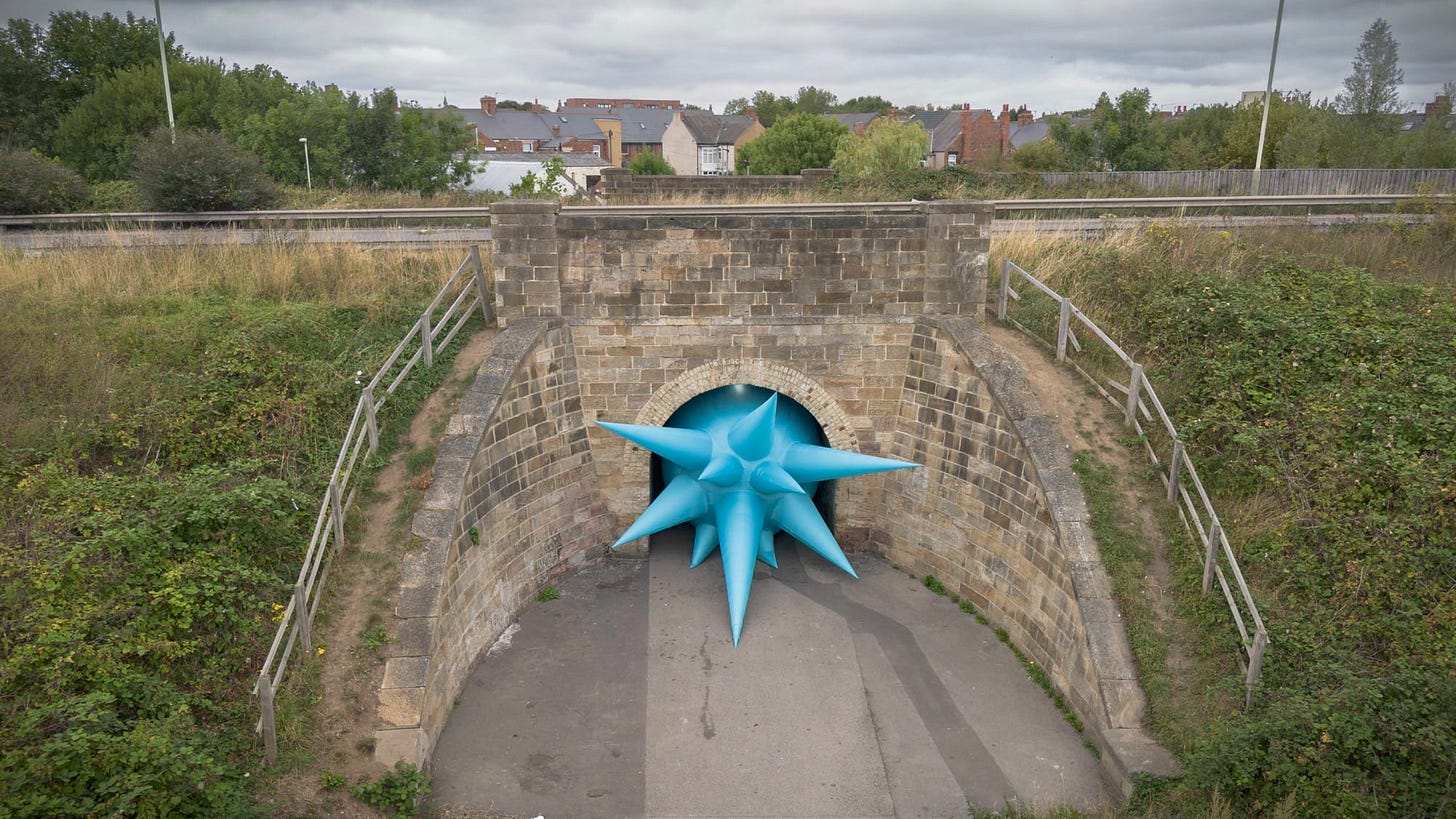 an aerial view of a large-scale installation by Steve Messam under an old stone bridge of a blue, spiked form that appears wedged in the archway under the bridge
