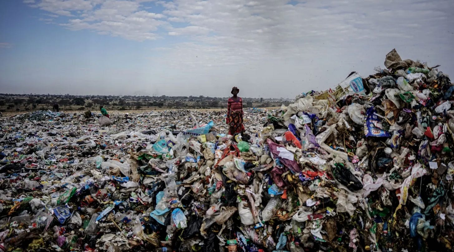 A person in a red patterned dress and hat stands on top of a vast open landfill site, surrounded by an enormous expanse of mixed plastic waste and rubbish stretching to the horizon, with sparse scrubland and a hazy sky in the background.