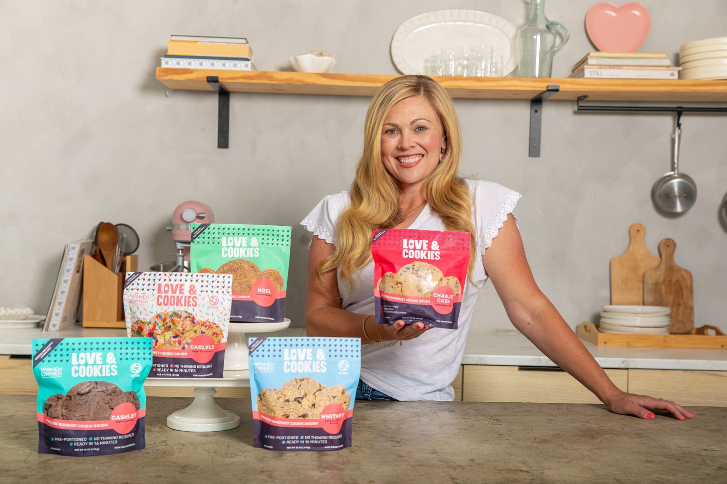 Ashley Cameron standing in a kitchen with exposed wooden shelves showing off her bags of Love & Cookies Ashley Cameron standing in a kitchen with exposed wooden shelves showing off her bags of Love & Cookies