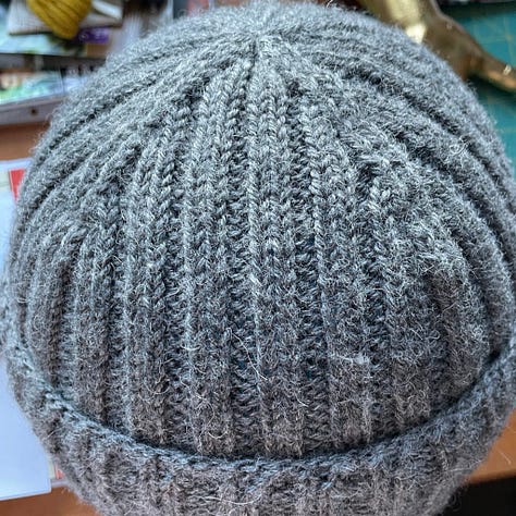 A hand knit hat in teal yarn; an identical hat in grey yarn; a woman with glasses wearing a grey watch cap.