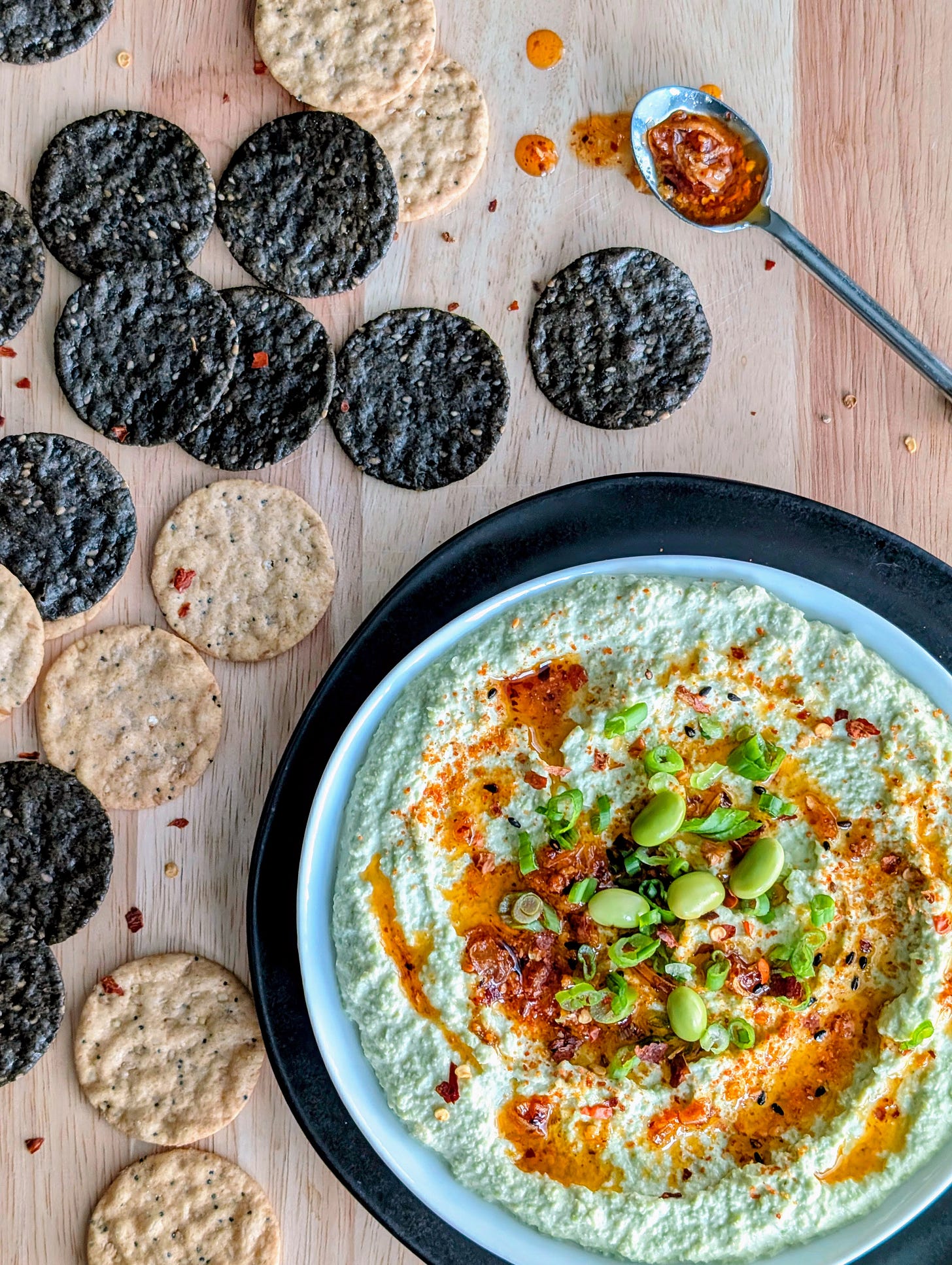 Bowl of creamy edamame dip topped with chili crisp, served with rice crackers on a wooden cutting board.