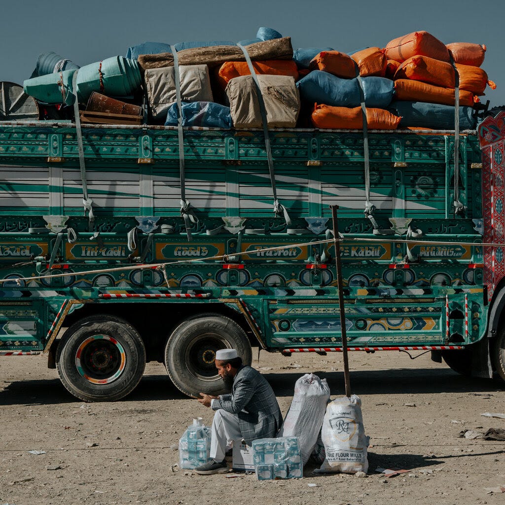 A person sits on dusty ground next to bottles of water and white sacks. A large green truck, brightly painted and heavily loaded with colorful bundles, is behind them.