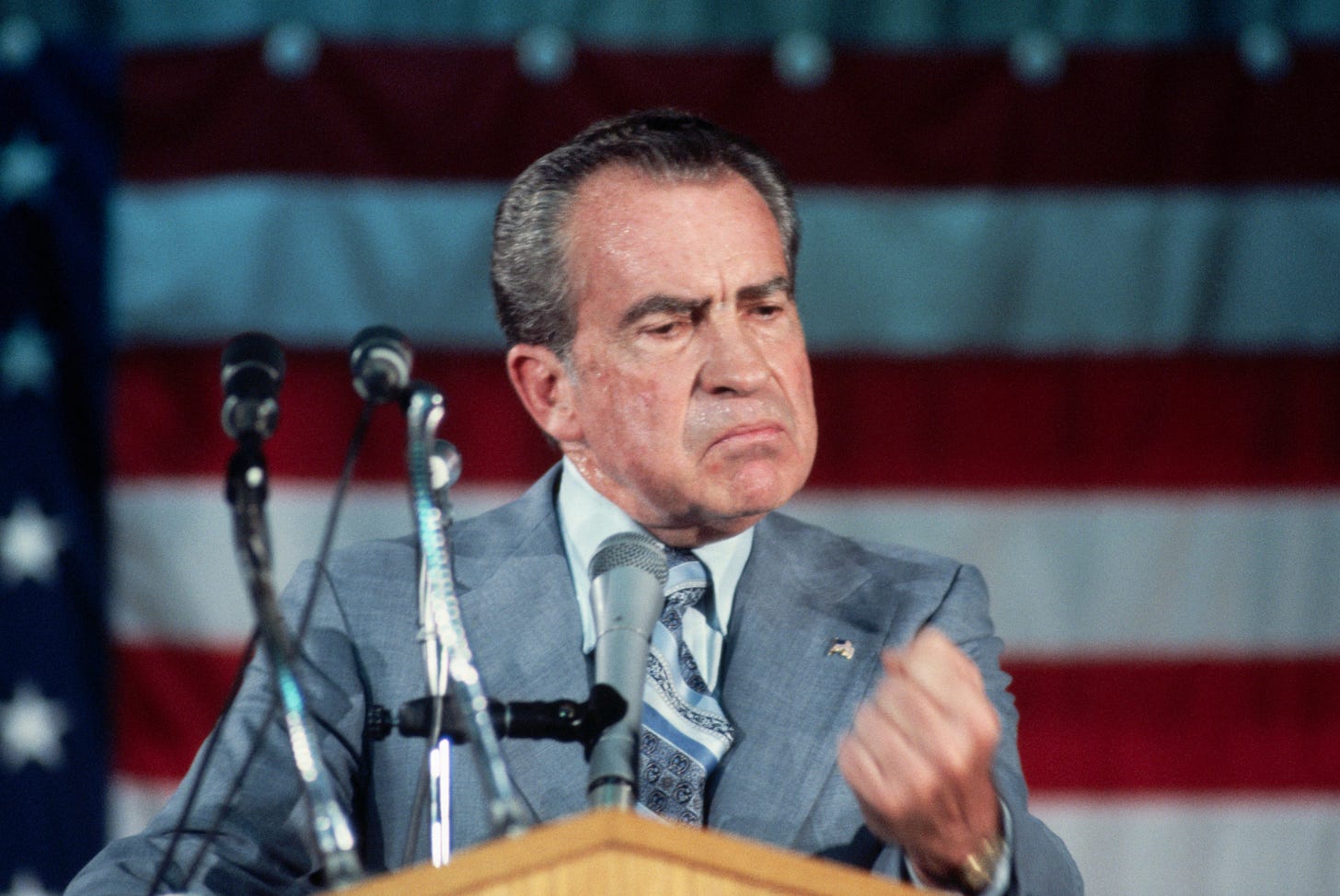 Richard Nixon, an older white man with swept back hair in a grey suit, stands behind a podium with microphones in front of an American flag. Richard Nixon, an older white man with swept back hair in a grey suit, stands behind a podium with microphones in front of an American flag.