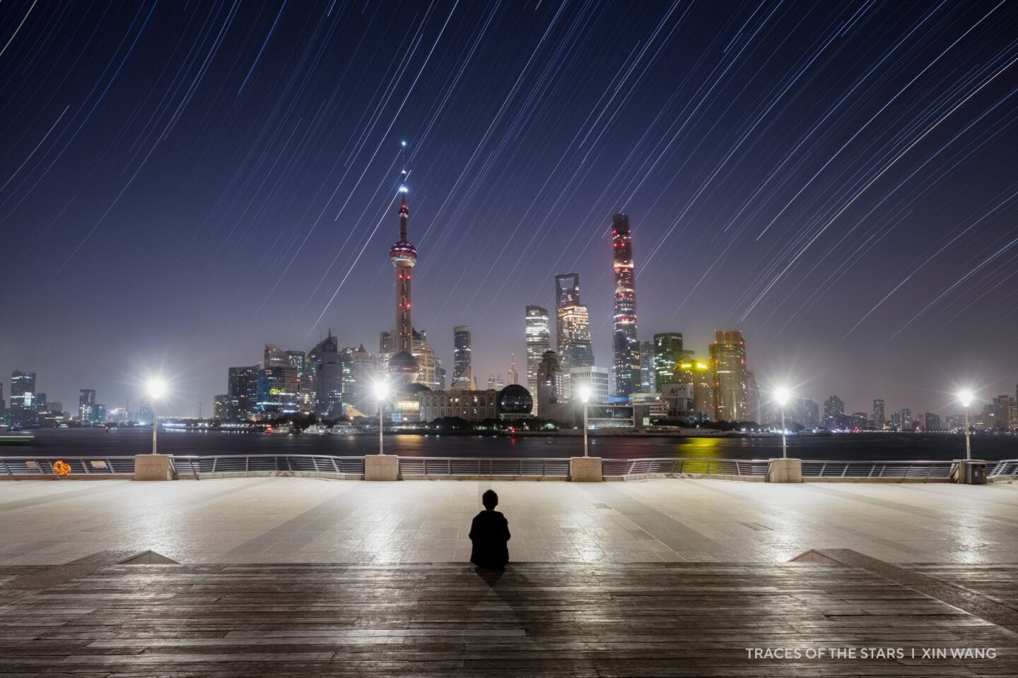 A person sits on a boardwalk facing a city skyline at night, with 2025 night sky images of star trails shimmering above the illuminated skyscrapers.