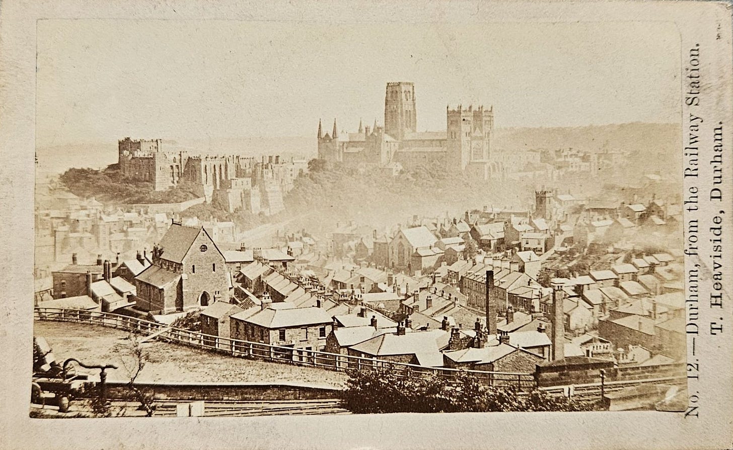 A vintage photograph depicting an aerial view of Durham, England, featuring rooftops of homes, a church, and the prominent Durham Cathedral in the background.