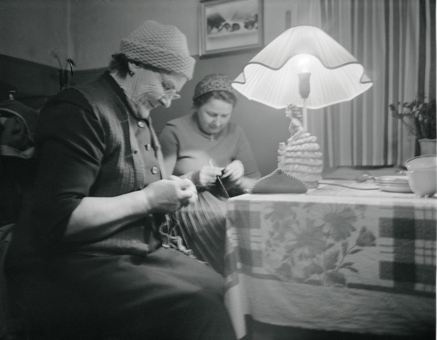 Two women sitting together indoors, both knitting, in a black-and-white photograph from 1960.